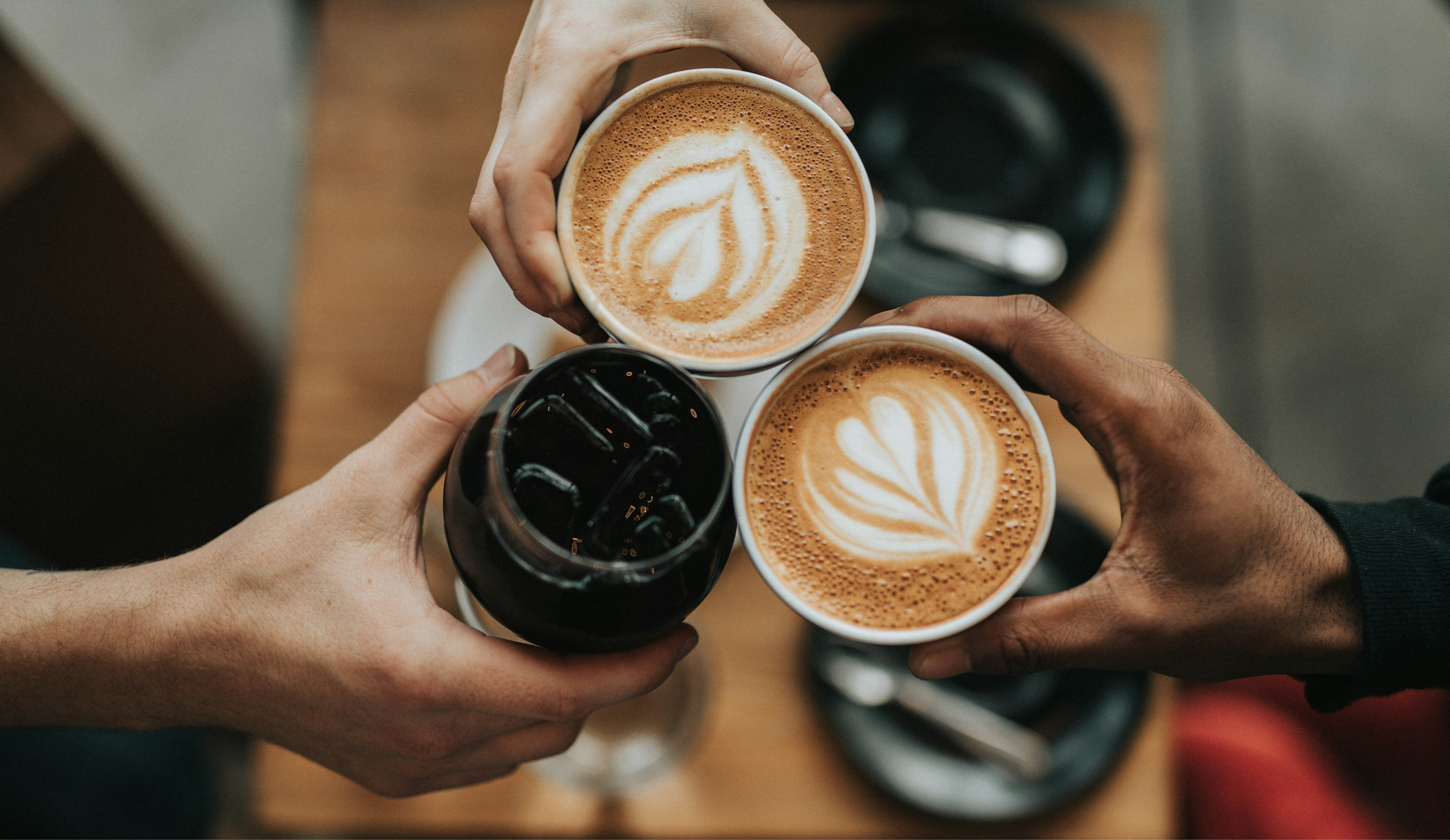 Three hands holding coffee cups with latte art on a wooden table.